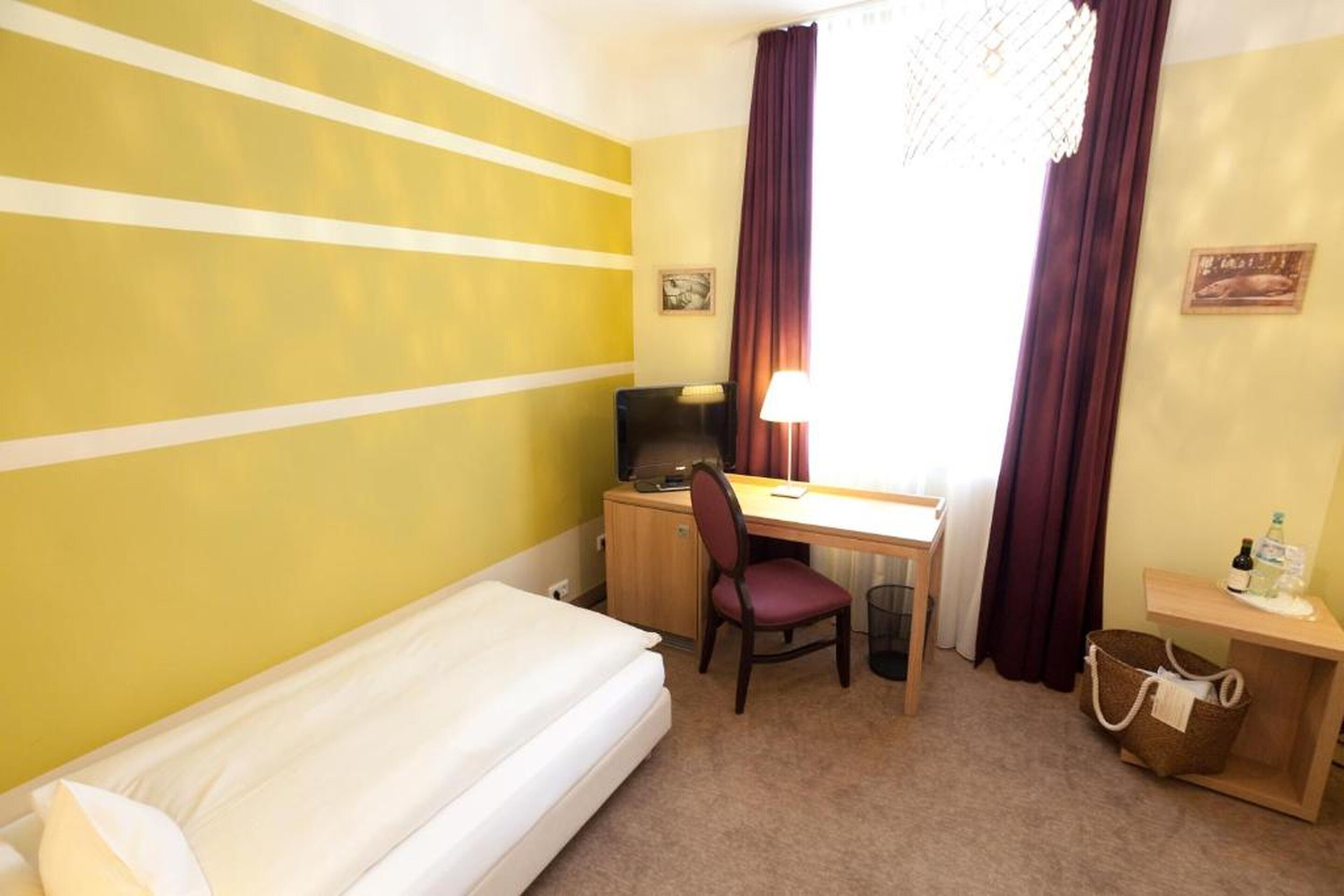 A hotel room with a bed, a desk, a chair, a lamp, and a small table with a bottle and a basket.