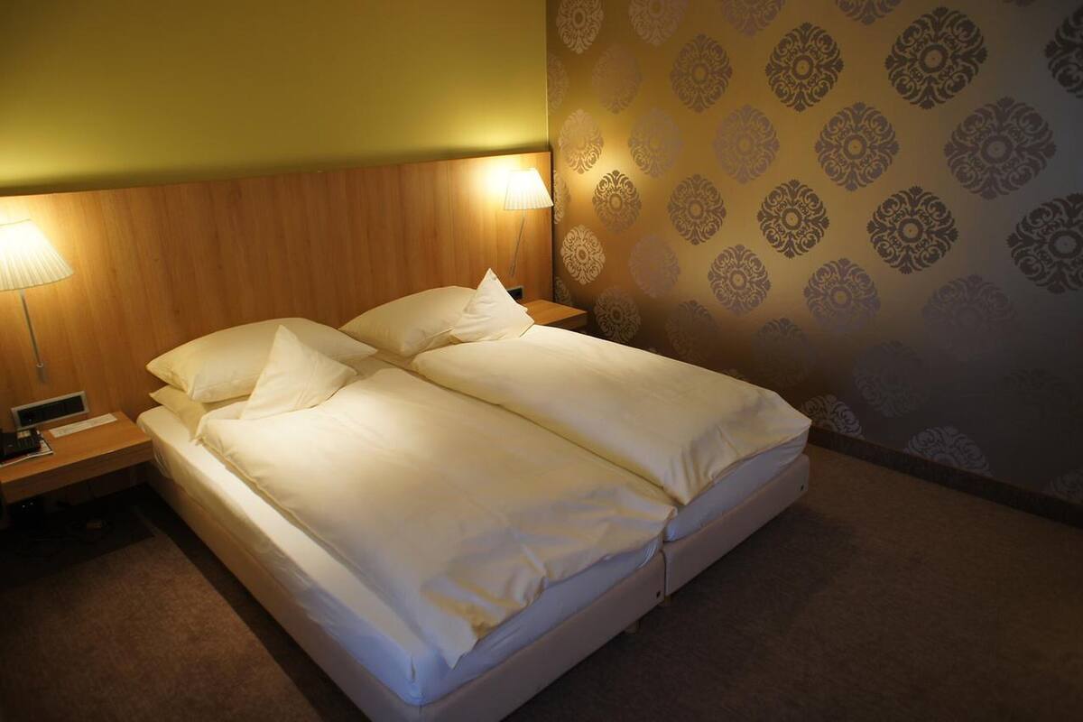 Ein Doppelbett mit weißen Leinen, ein Holz-Kopfteil, eine Nachttischlampe und eine Wand mit Musterpapier.