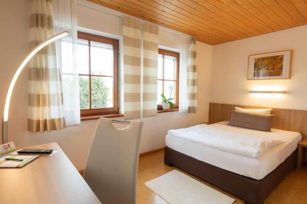 A hotel room with a bed, a desk, a chair, two windows with curtains, a wall-mounted lamp, and a framed picture.