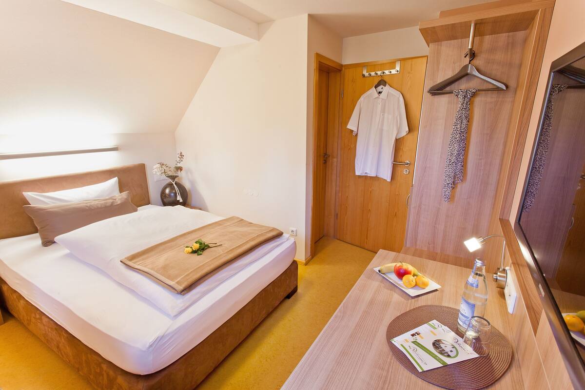 A hotel room with a bed, a wooden bedside table with a tray of fruit, a bottle of water, and a magazine.