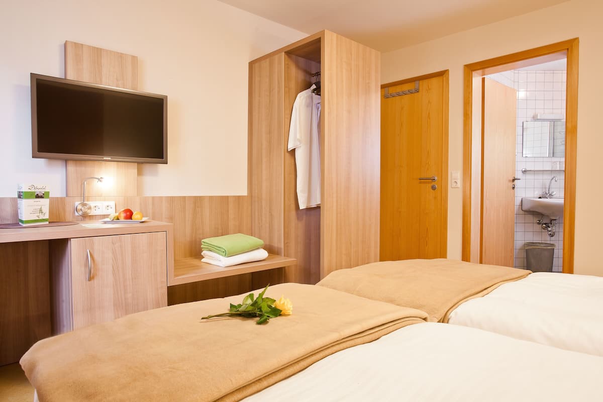A hotel room with two beds, a TV mounted on the wall, a wooden wardrobe, a small desk with a mirror, and a bathroom visible through an open door.