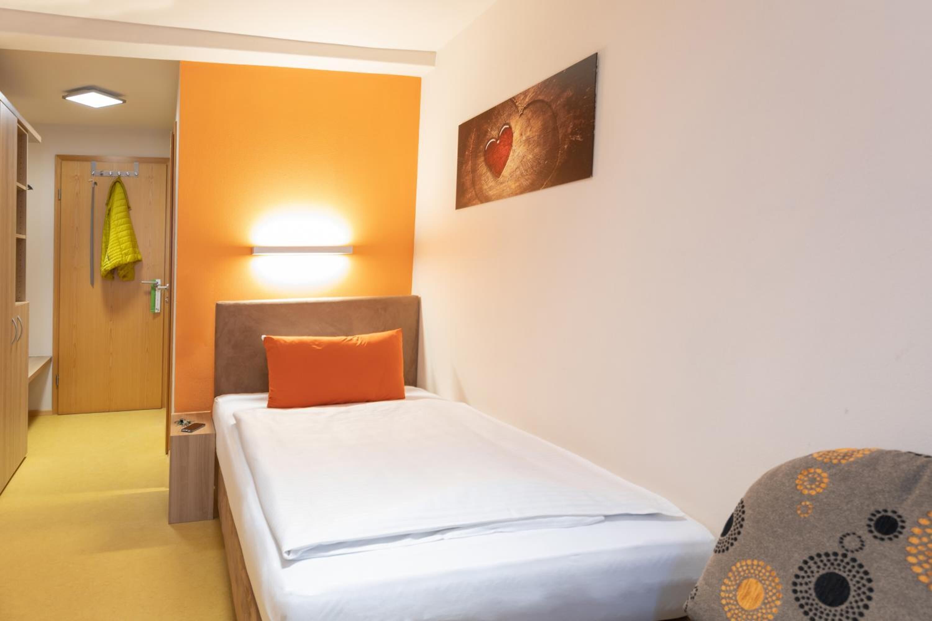 Habitación de hotel con una cama, una mesita de noche, una lámpara, una puerta con un abrigo colgado y un cuadro en la pared.