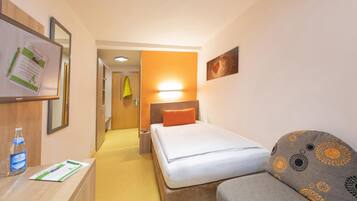Quarto de hotel com cama, sofá, escrivaninha com uma garrafa e um menu, espelho e um quadro na parede.