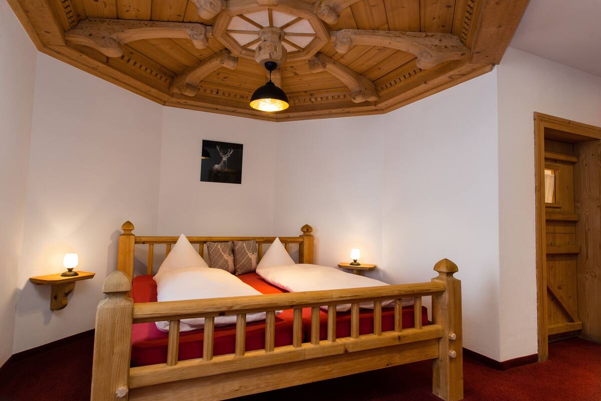 Uma cama de madeira com colcha vermelha em quarto com paredes brancas e teto de madeira.