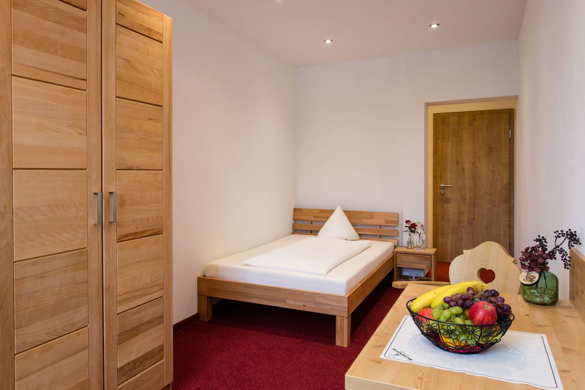 Quarto com cama de madeira, tapete vermelho, mesa de cabeceira de madeira com um vaso de flores e uma cesta de frutas sobre uma mesa.