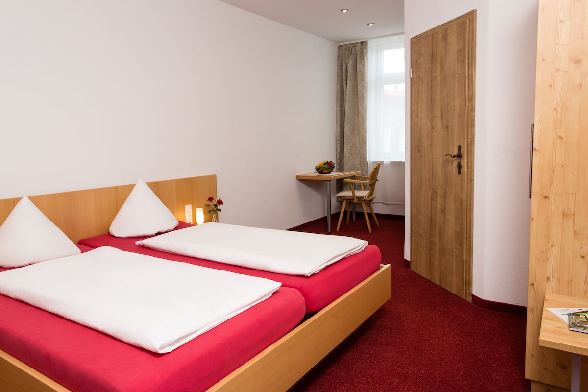 Quarto de hotel com duas camas, uma escrivaninha de madeira, uma cadeira e uma porta.