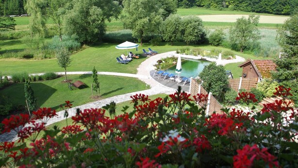 Un espace piscine agrémenté de chaises longues et d’une fontaine, entouré d’une pelouse bien entretenue et d’arbres.