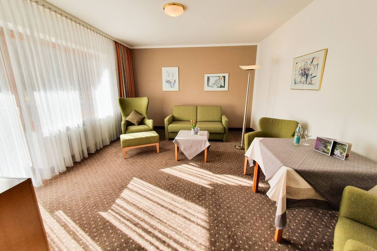 Une chambre d’hôtel avec un canapé vert, un fauteuil vert, une petite table recouverte d’une nappe blanche et un lampadaire. Deux tableaux encadrés ornent le mur, et une fenêtre est agrémentée de rideaux légers.