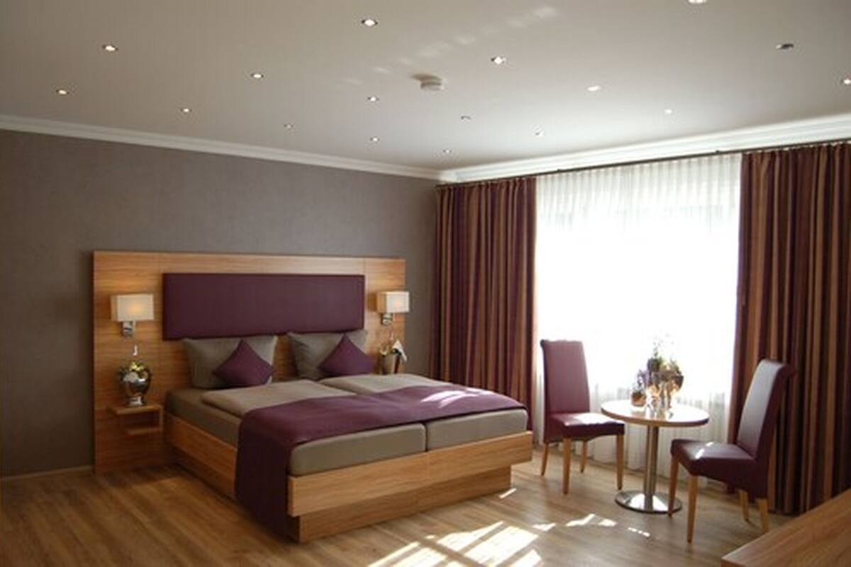 A modern hotel room with a large bed, a small table, two chairs, and a large window with curtains.