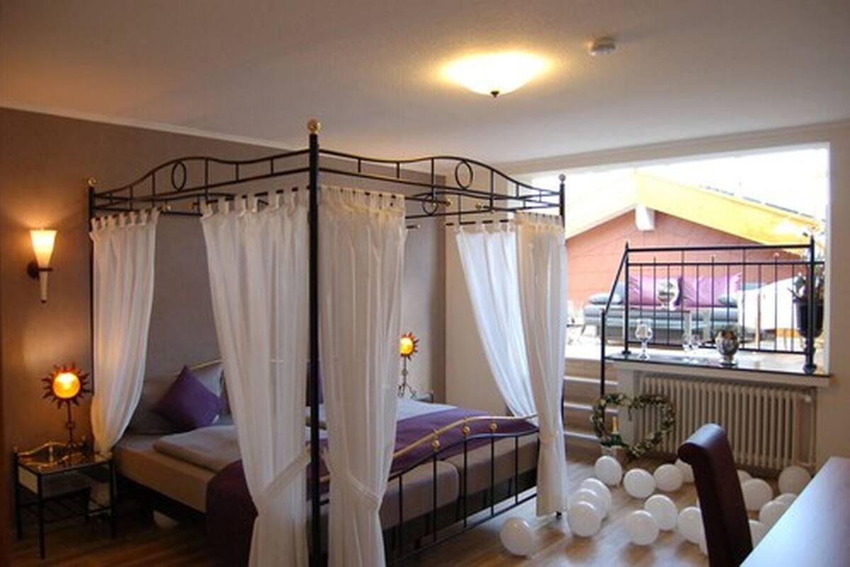 A bedroom with a four-poster bed, white curtains, a bedside lamp, and a view of the outside.