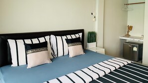 A neatly arranged bedroom with a bed, pillows, a bedside table with a plant, a mirror, and a small refrigerator. - Lom La Lanta Hotel (Ko Lanta)