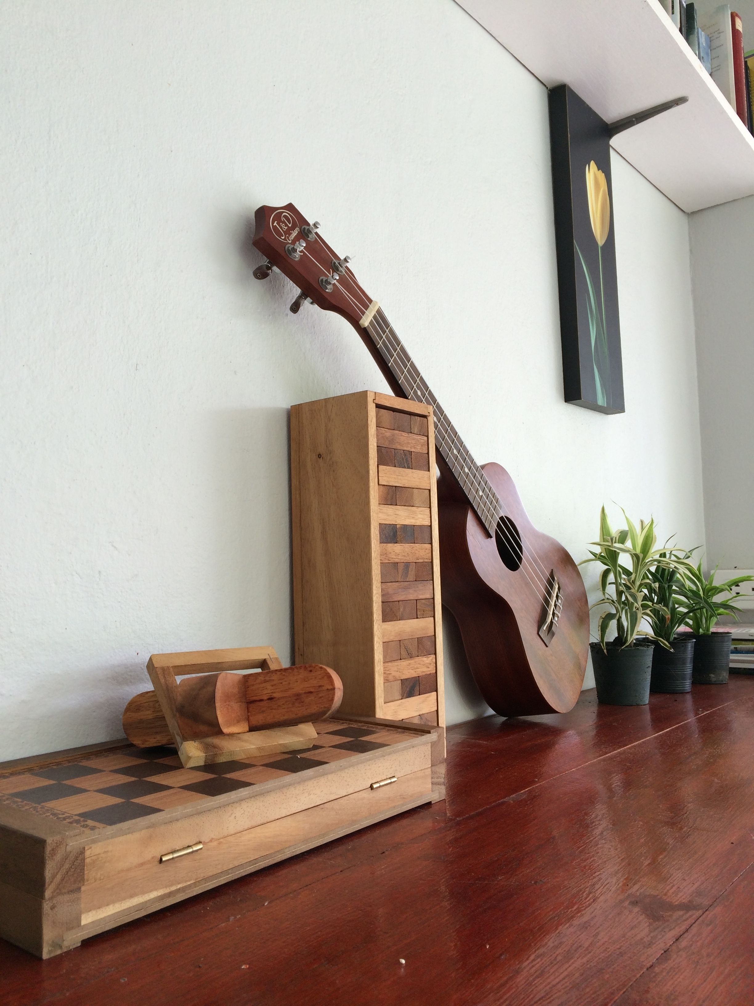 A wooden guitar leaning against a wall, a small wooden cabinet, a chessboard, and potted plants on a wooden floor.