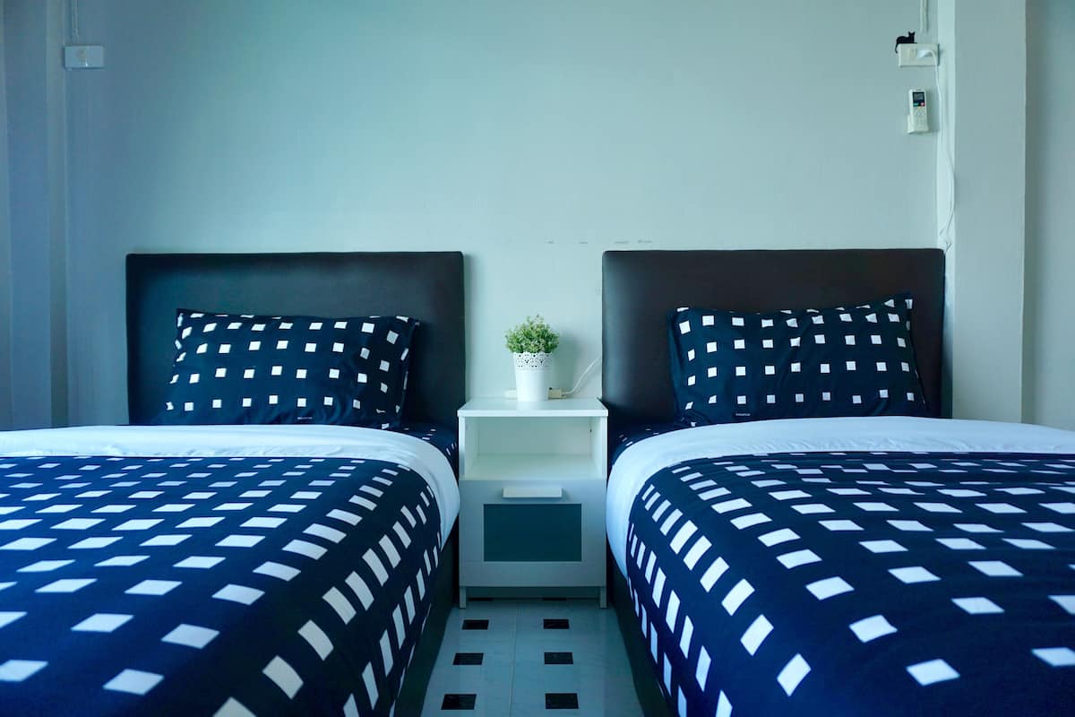 A hotel room with two beds, each with a dark blue and white checkered bedspread, a white nightstand with a small plant, and a white wall with a thermostat and a power outlet.