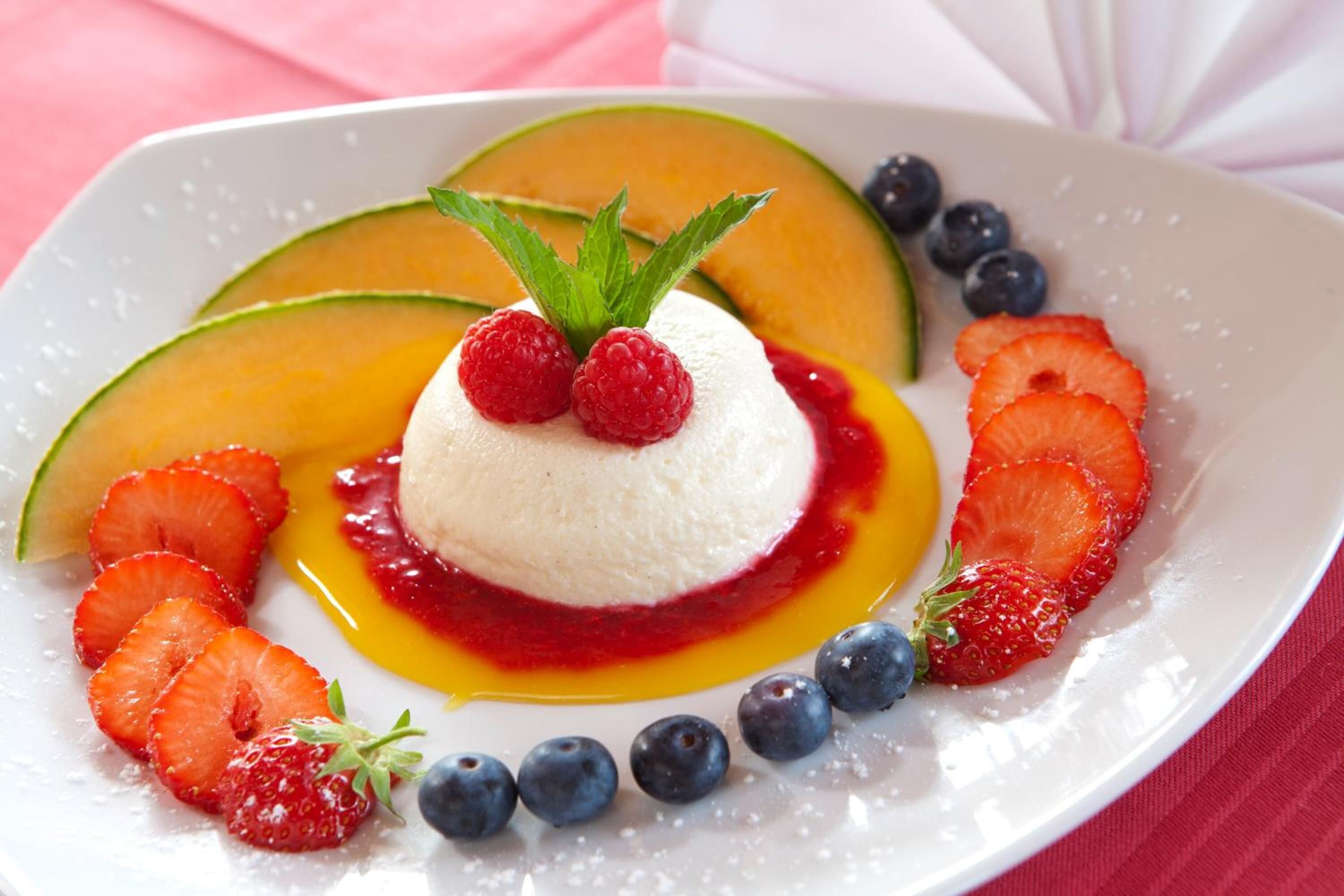A dessert with a scoop of ice cream topped with raspberries and mint, surrounded by sliced strawberries and blueberries, with a slice of melon and a drizzle of sauce on a white plate.