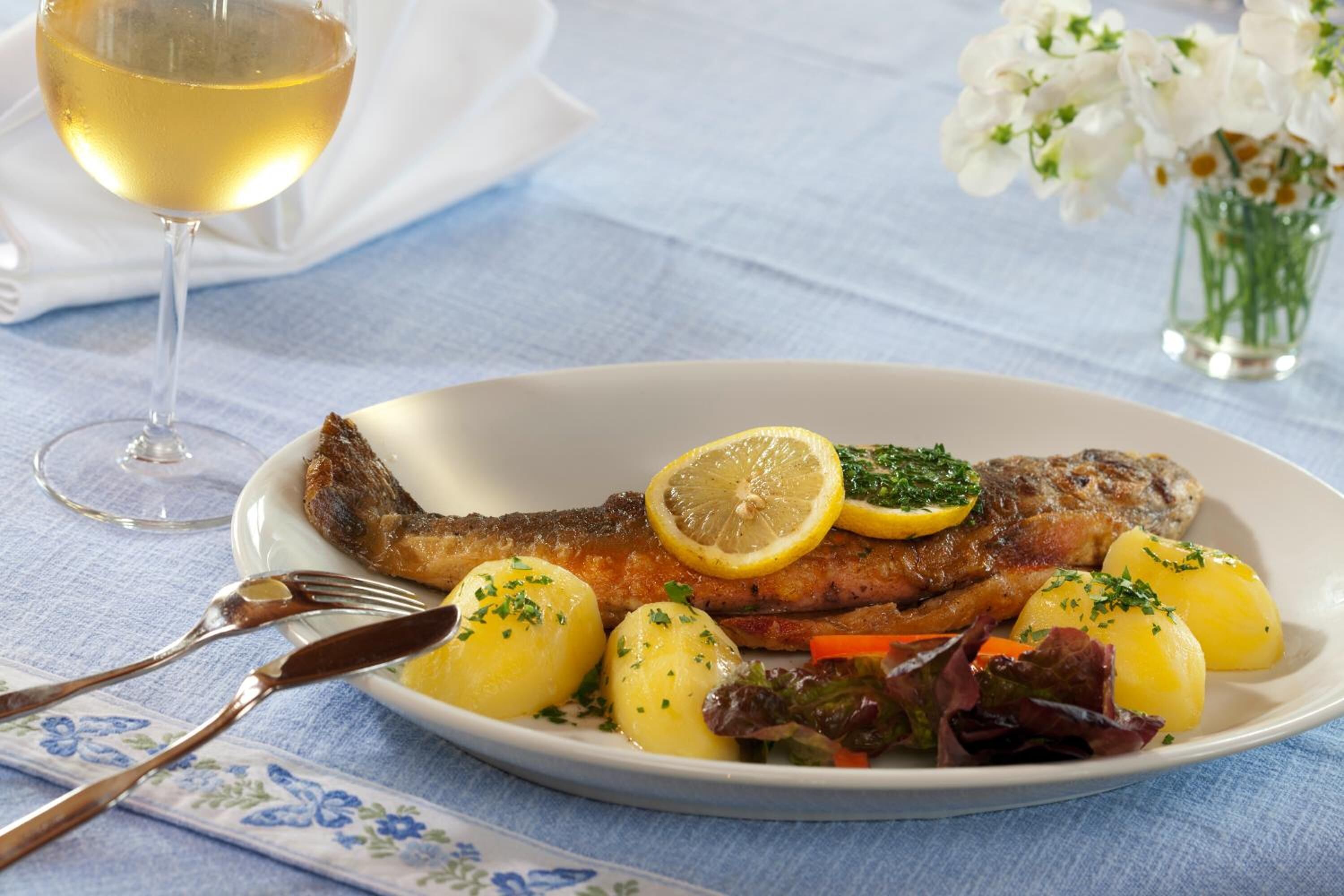 A plate of food with a fish, lemon slices, and potatoes, accompanied by a glass of white wine.