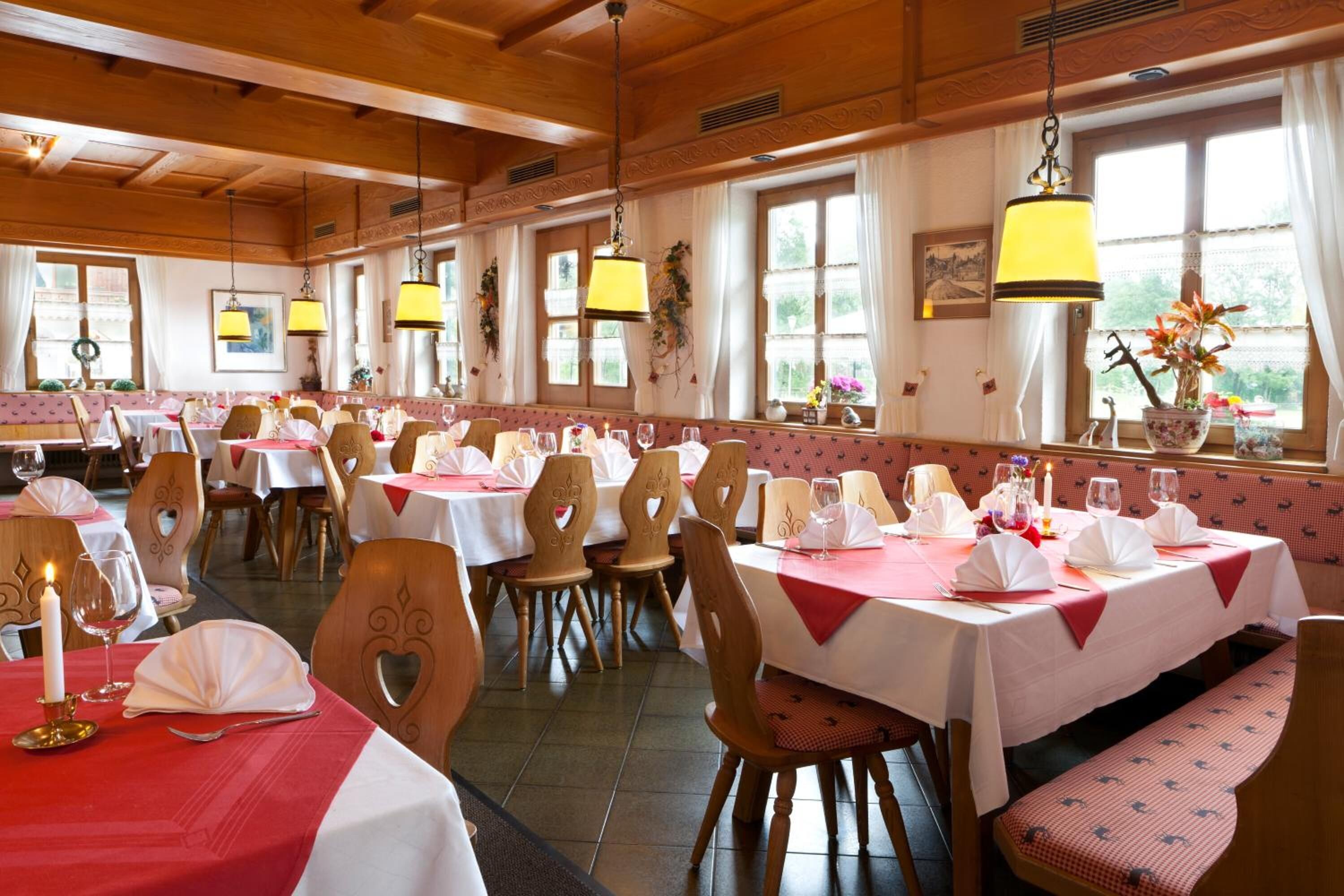 A restaurant with wooden interiors, multiple tables set with white tablecloths and red napkins, and large windows with curtains.