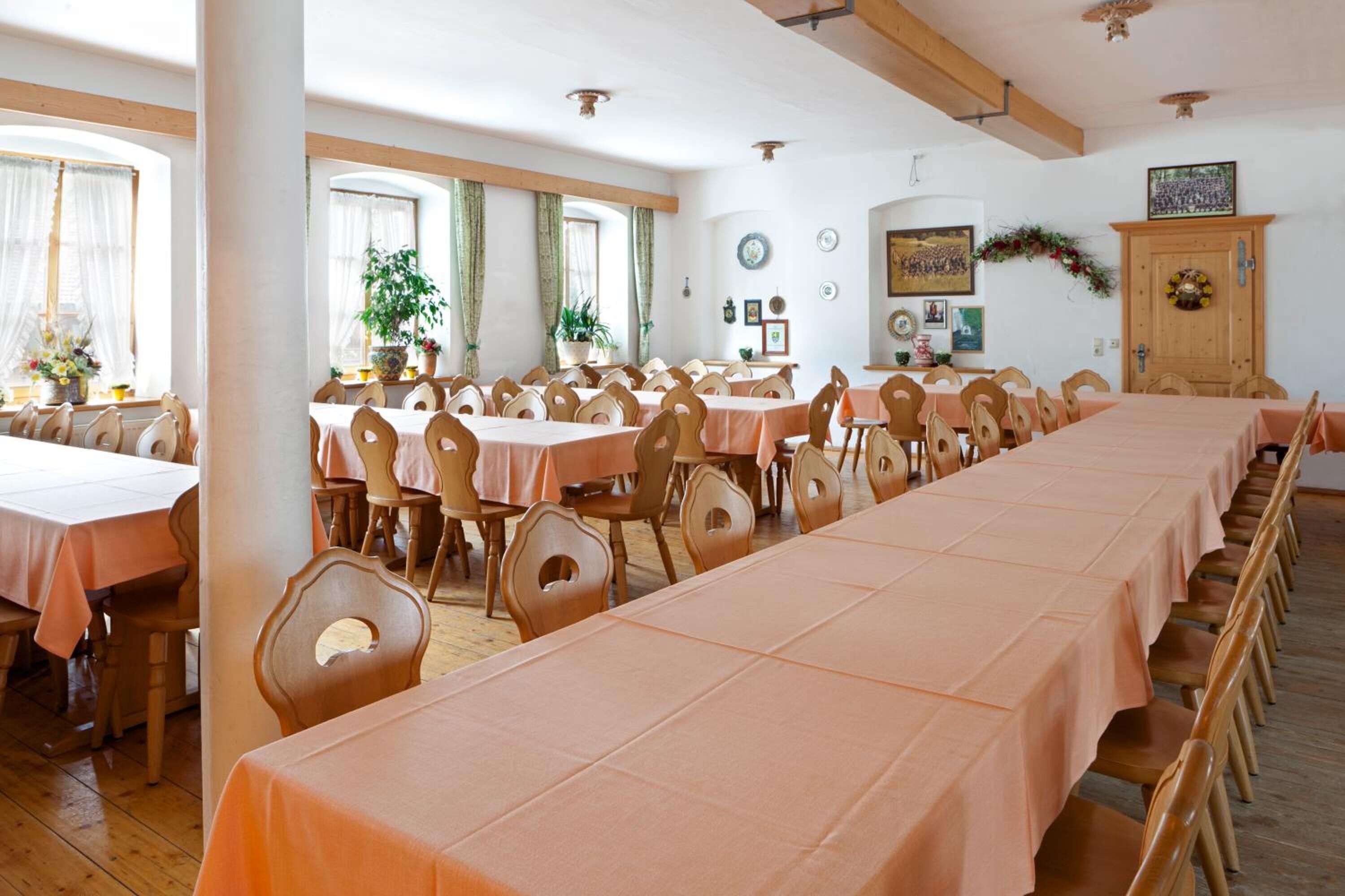 A large dining hall with multiple tables and chairs, wooden beams on the ceiling, and large windows with curtains.