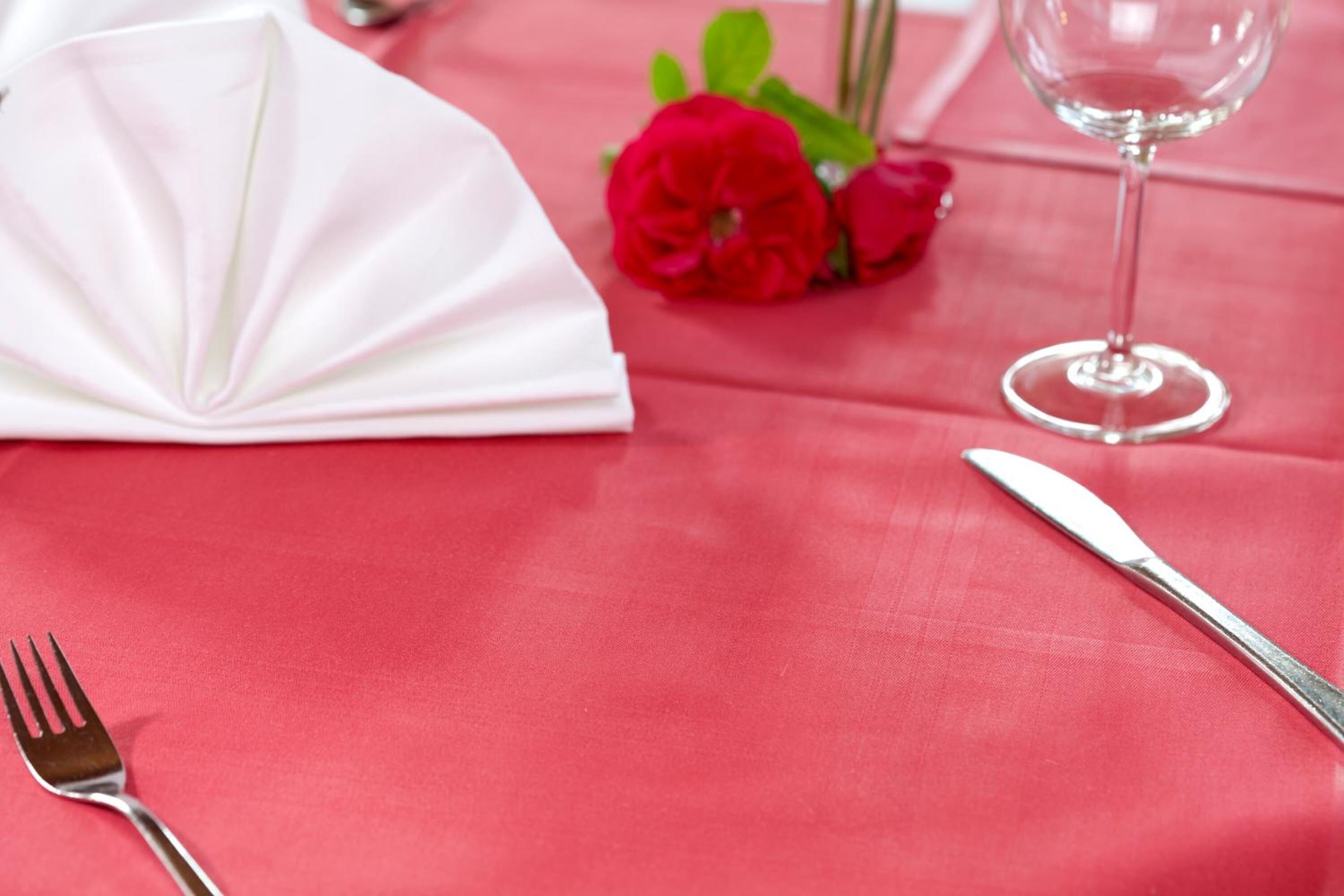 Une table recouverte d’une nappe rouge, avec une serviette blanche, une fourchette, un couteau, un verre d’eau et une fleur rouge.