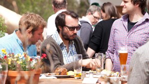 A group of people dining outdoors at a wooden table with potted plants and clear glasses of beer.