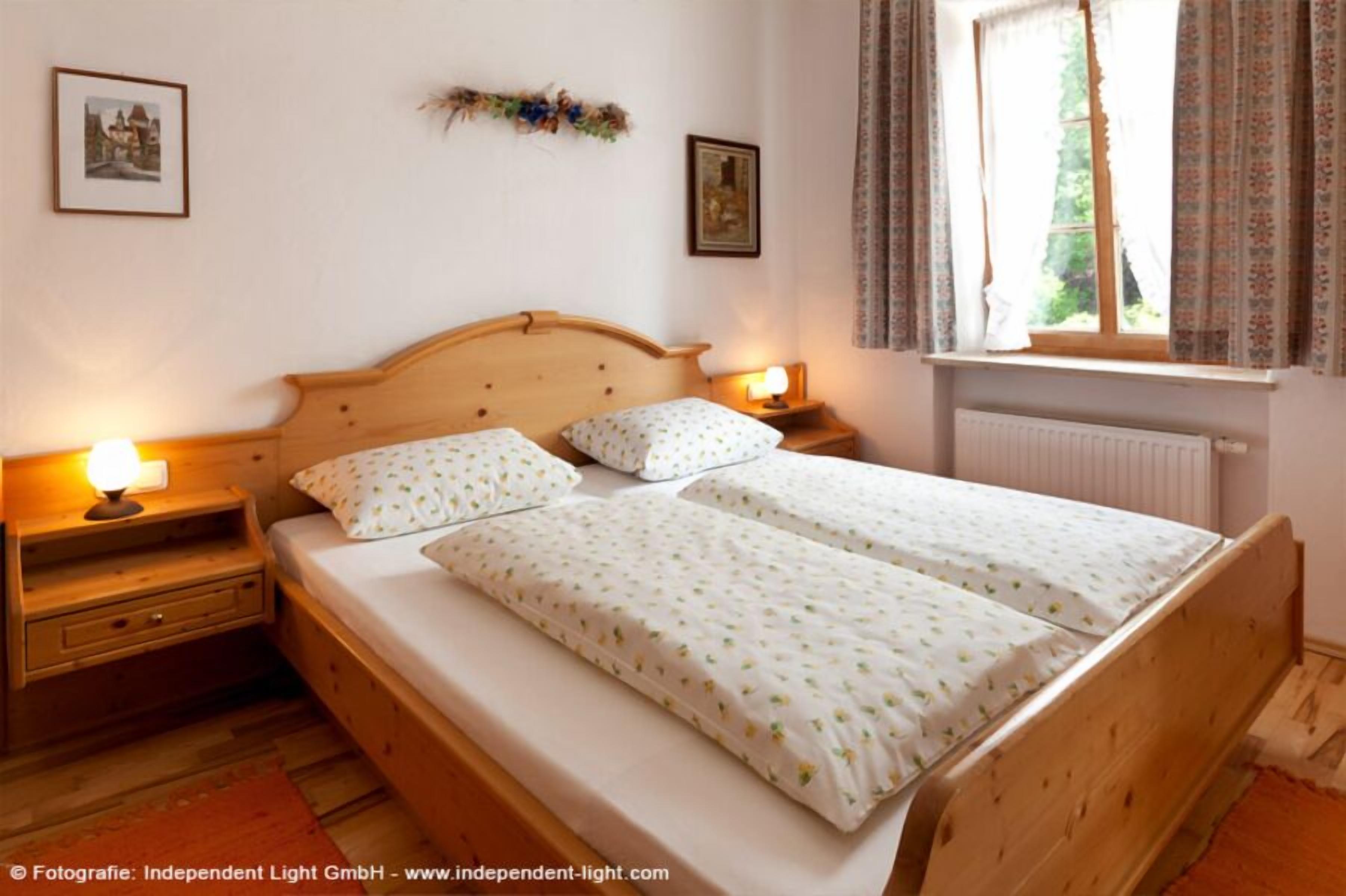 Cama doble con dos almohadas, cabecero de madera, mesitas de noche con lámparas y una ventana con cortinas.