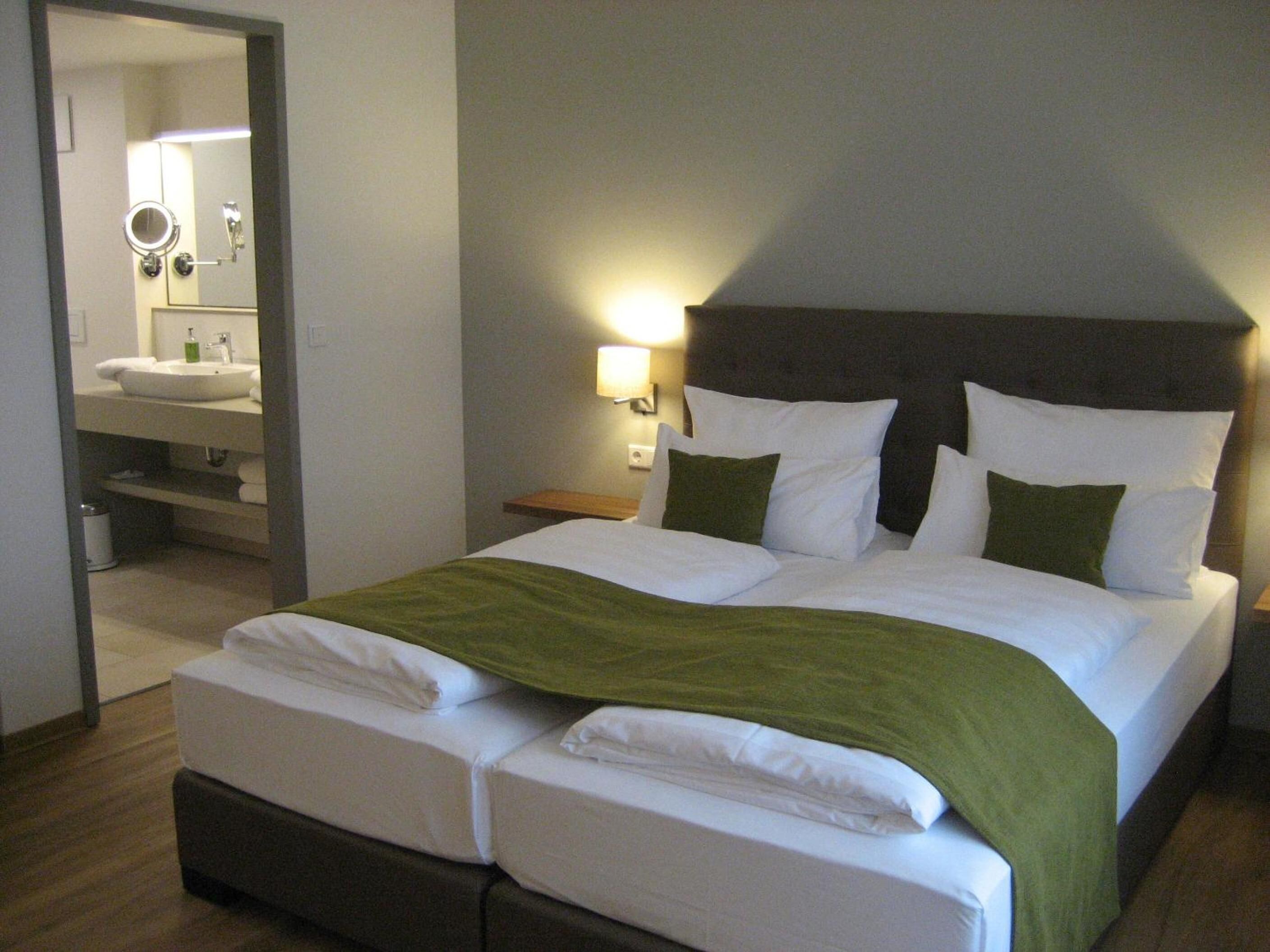 A hotel room with a double bed, a nightstand with a lamp, a bathroom with a sink and mirror, and a green throw blanket on the bed.