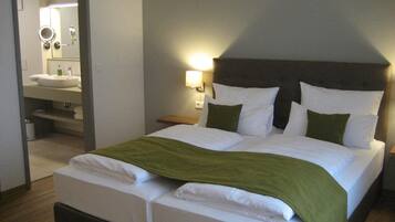 A hotel room with a double bed, a nightstand with a lamp, a bathroom with a sink and mirror, and a green throw blanket on the bed.