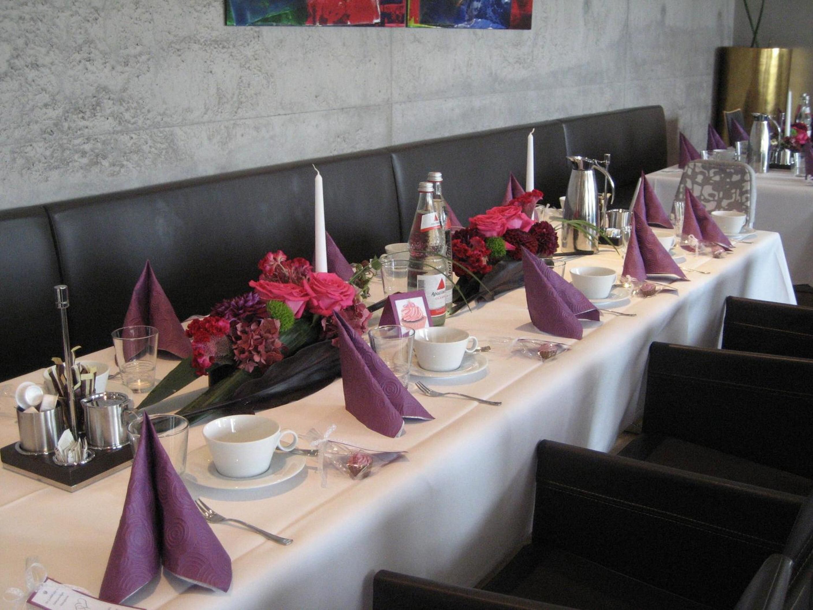 Ein langer Esstisch, gedeckt mit einer weißen Tischdecke, violetten Servietten und floralen Tischdekorationen.