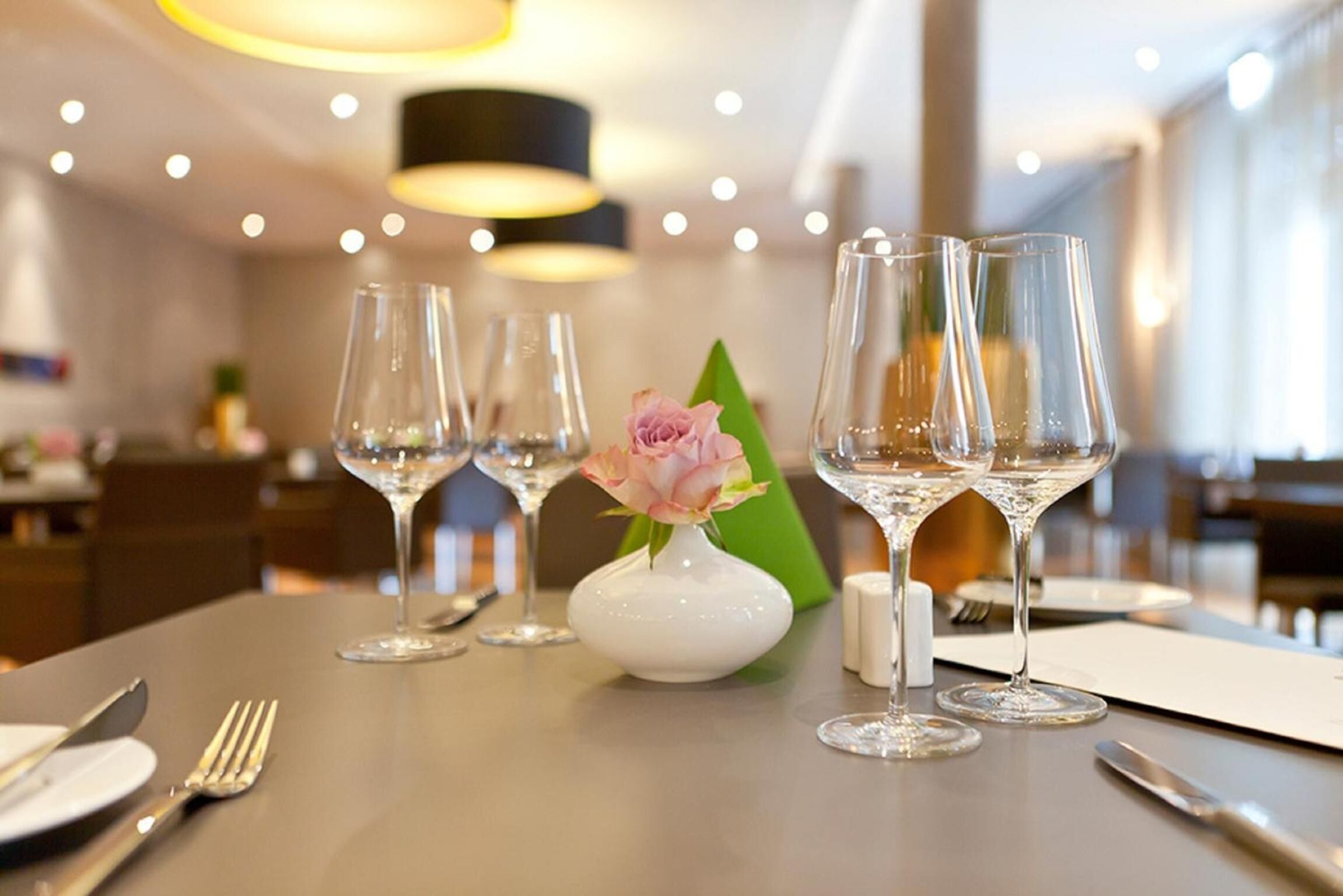Una mesa de comedor con cuatro copas de vino, un jarrón blanco con una flor rosa y un portavasos verde.