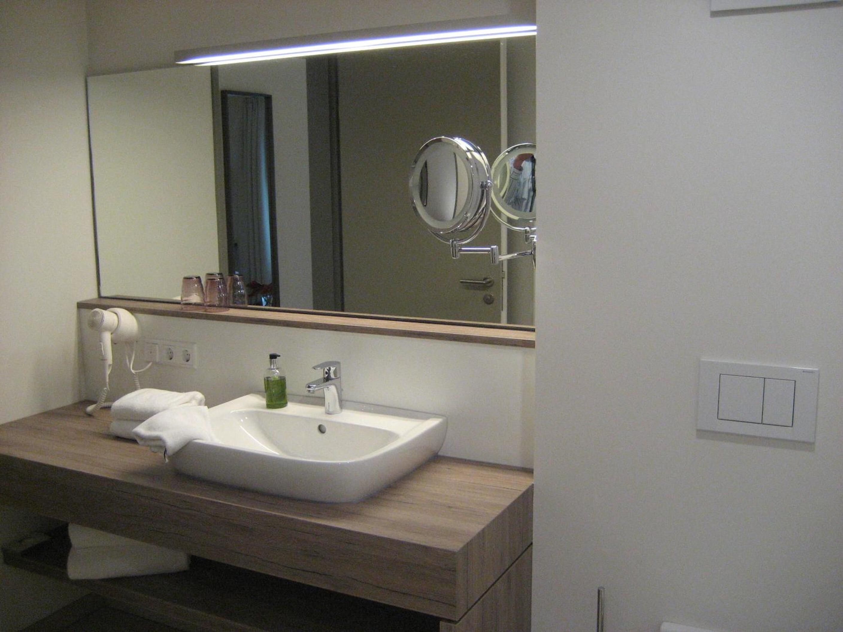 Un baño con lavamanos blanco, un espejo y un secador de pelo sobre una cómoda de madera.