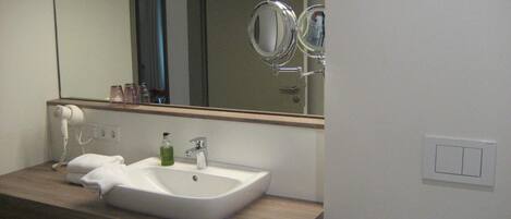 Un baño con lavamanos blanco, un espejo y un secador de pelo sobre una cómoda de madera.