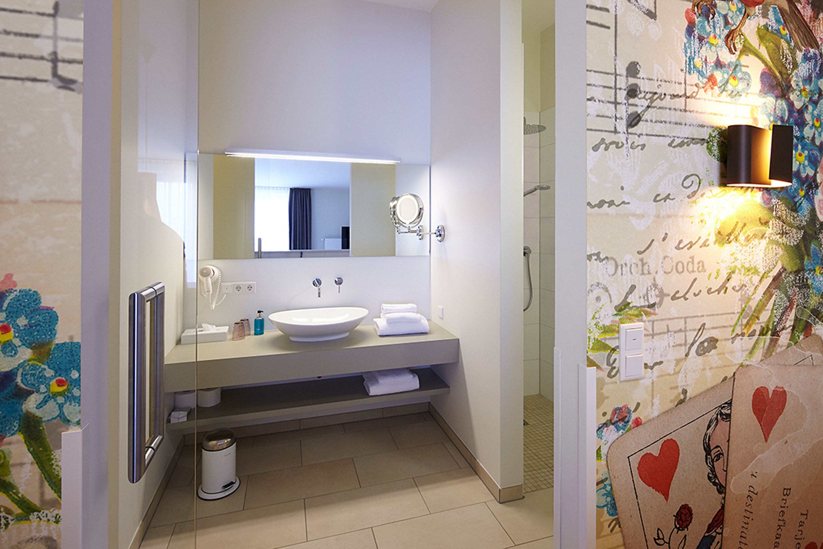 Une salle de bain avec un lavabo blanc, un miroir et un porte-serviettes. On peut voir une poubelle et une zone douche.