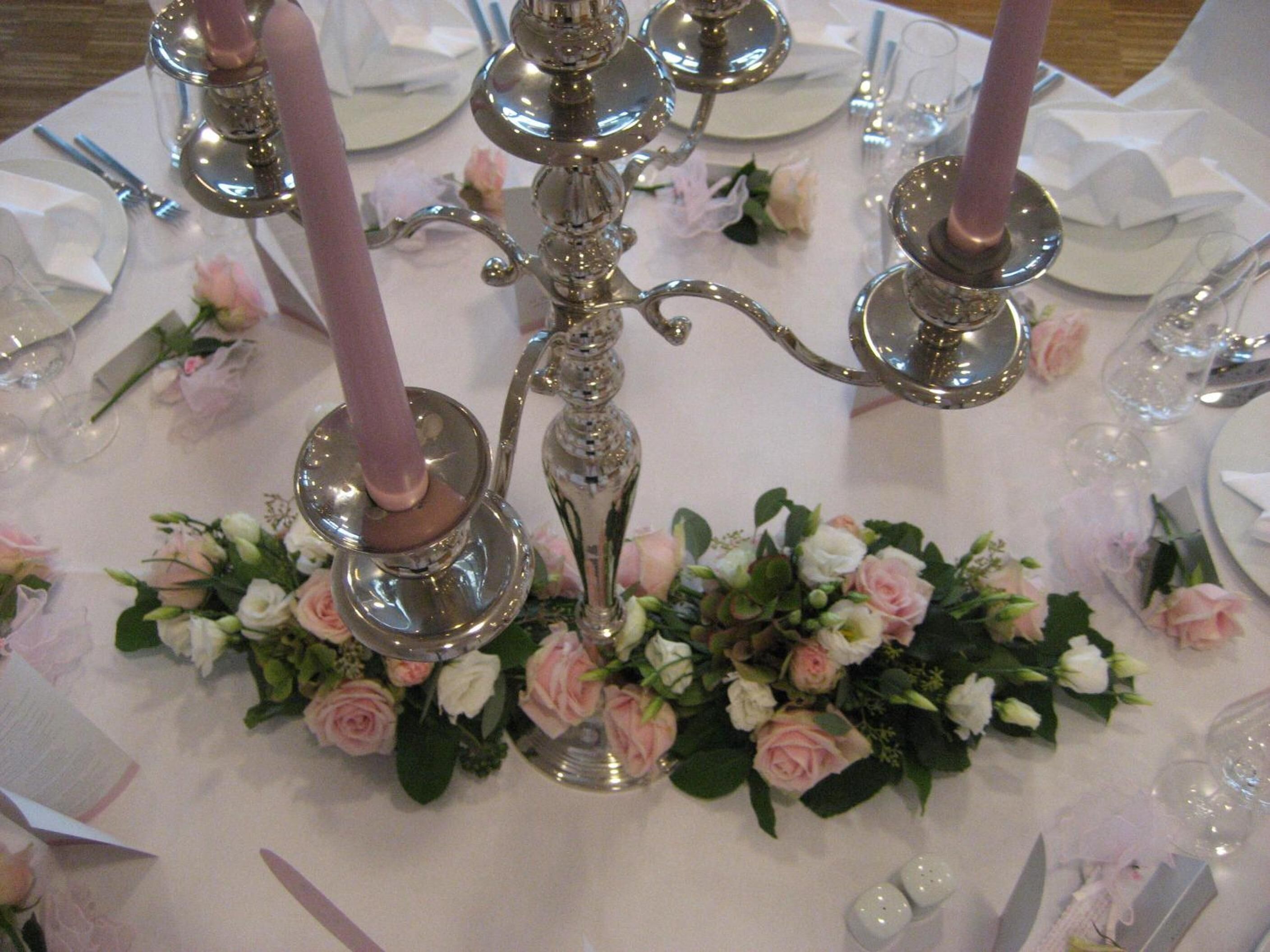 Une table dressée pour un événement formel, avec des couverts en argent, des assiettes et des bougies roses.