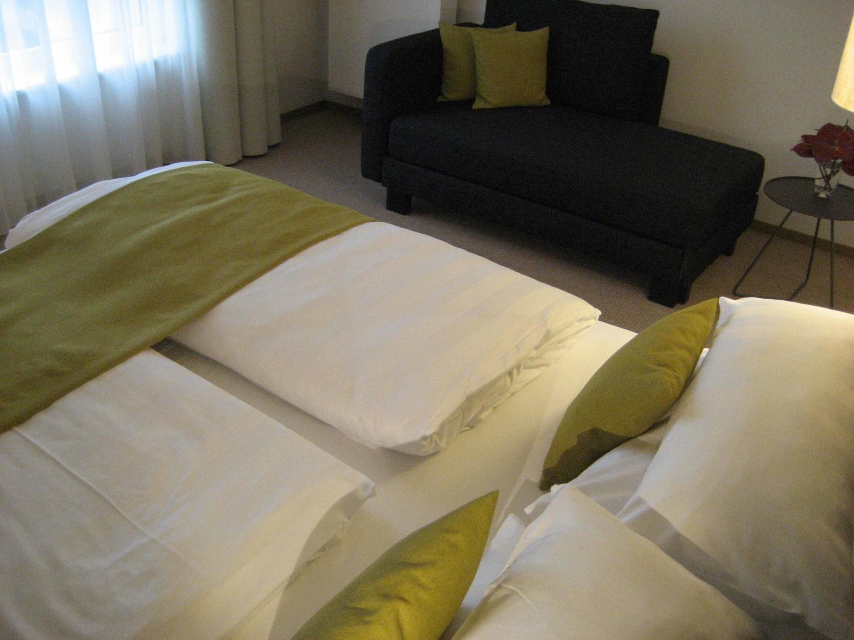 Habitación de hotel con dos camas, un sofá oscuro, una mesita redonda con un florero y una ventana con cortinas.