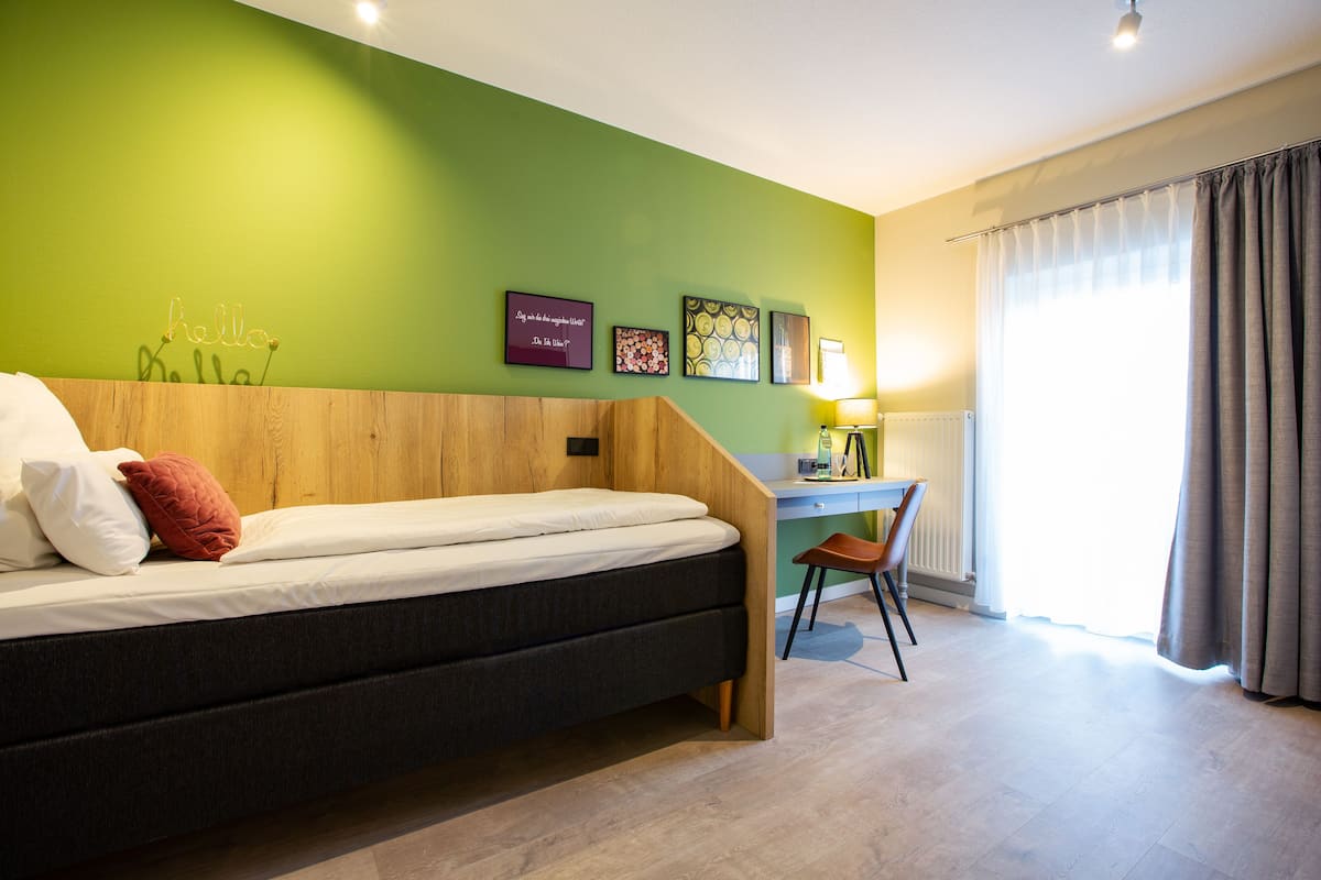 Une pièce comprenant un lit, un bureau et une chaise. Les murs sont ornés de tableaux et il y a une fenêtre avec des rideaux.