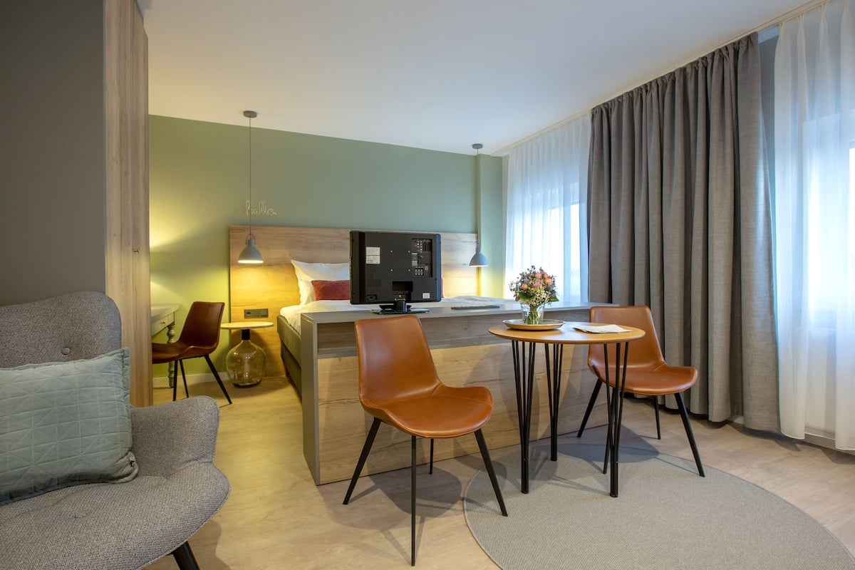 Une chambre d’hôtel avec un lit, un bureau, deux chaises, une lampe et un vase avec des fleurs.