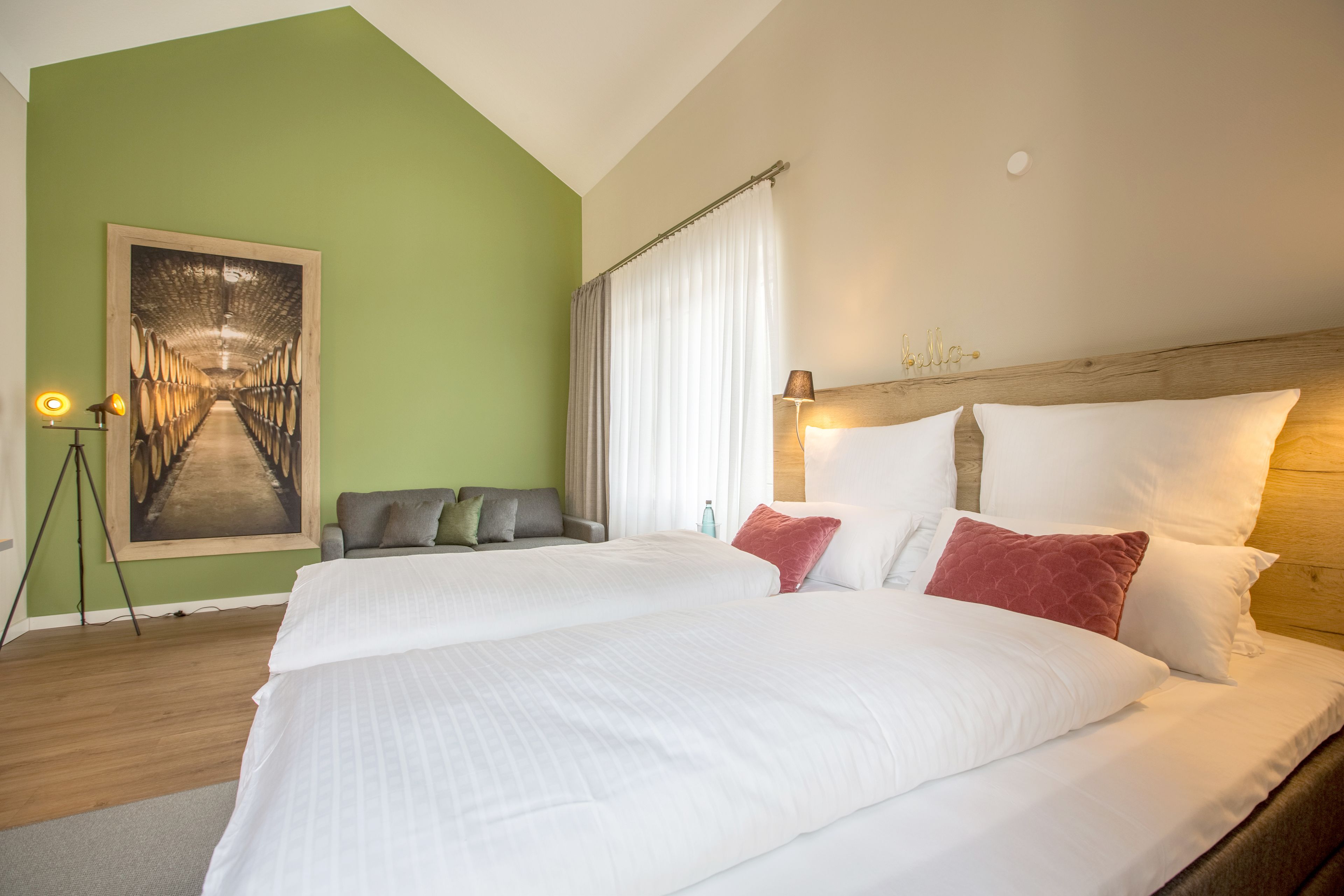 Una cama doble con sábanas blancas y almohadas rojas, un cabecero de madera, una pared verde, un cuadro enmarcado de un túnel y un sofá gris.