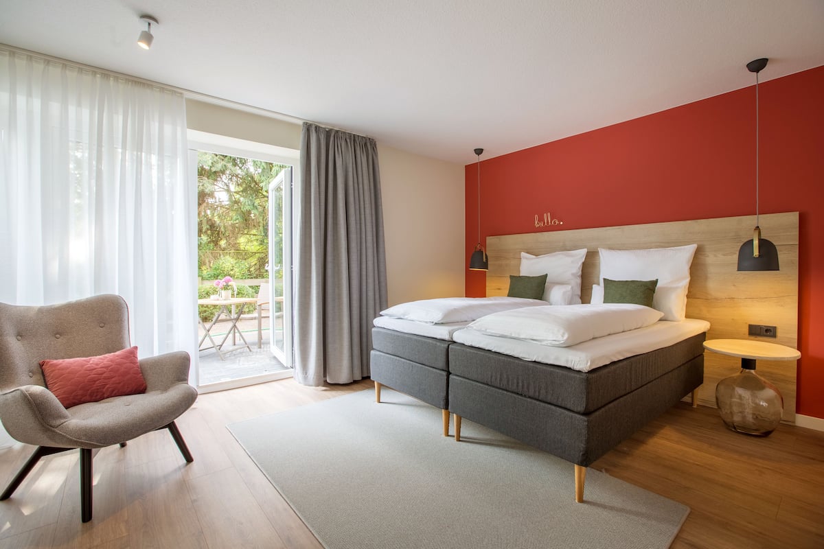 Une chambre moderne avec un lit gris, un mur d’accent rouge et une grande fenêtre avec des rideaux.