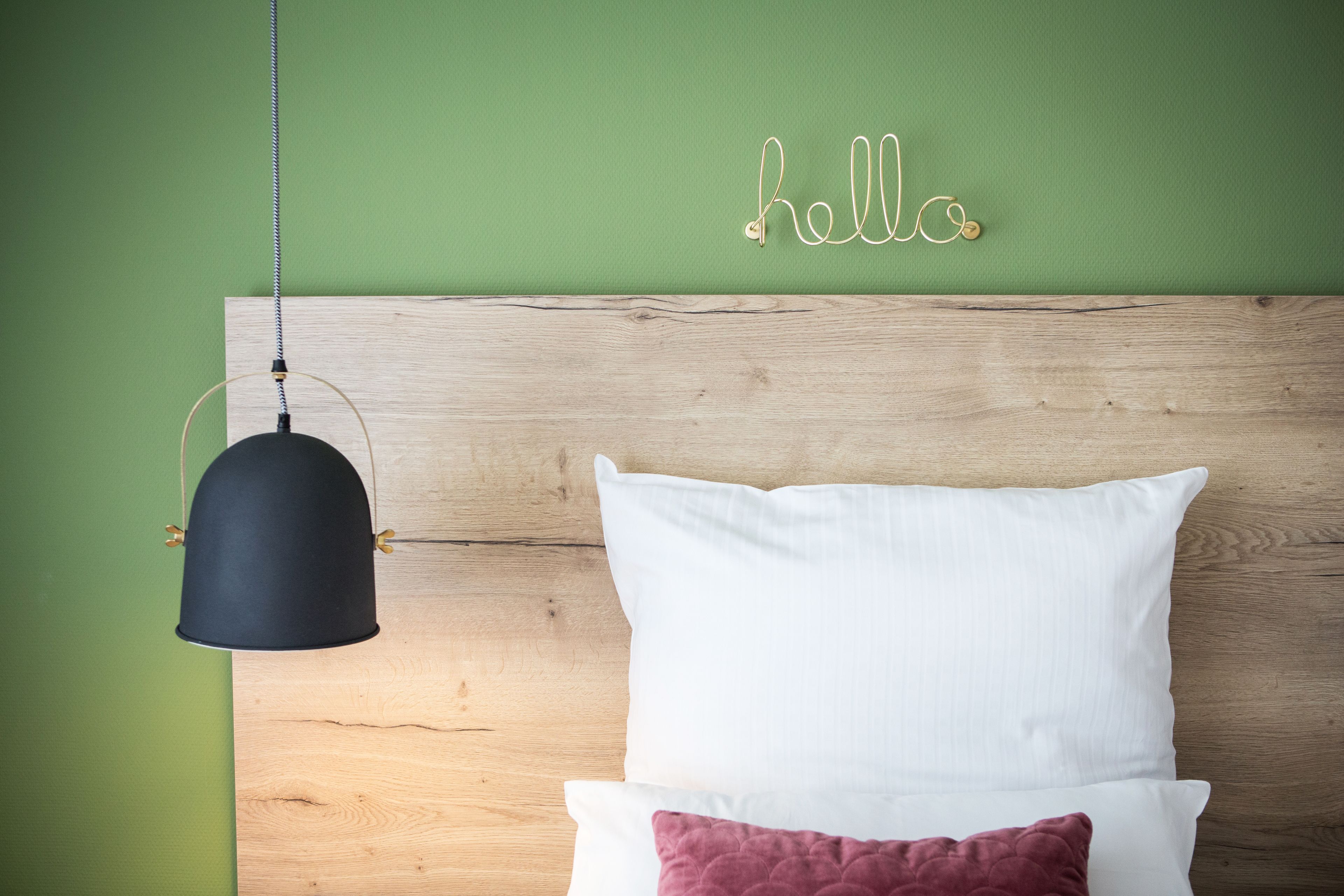 A bed with white and maroon pillows, a black hanging lamp, and a wooden headboard against a green wall with a 'hello' sign.