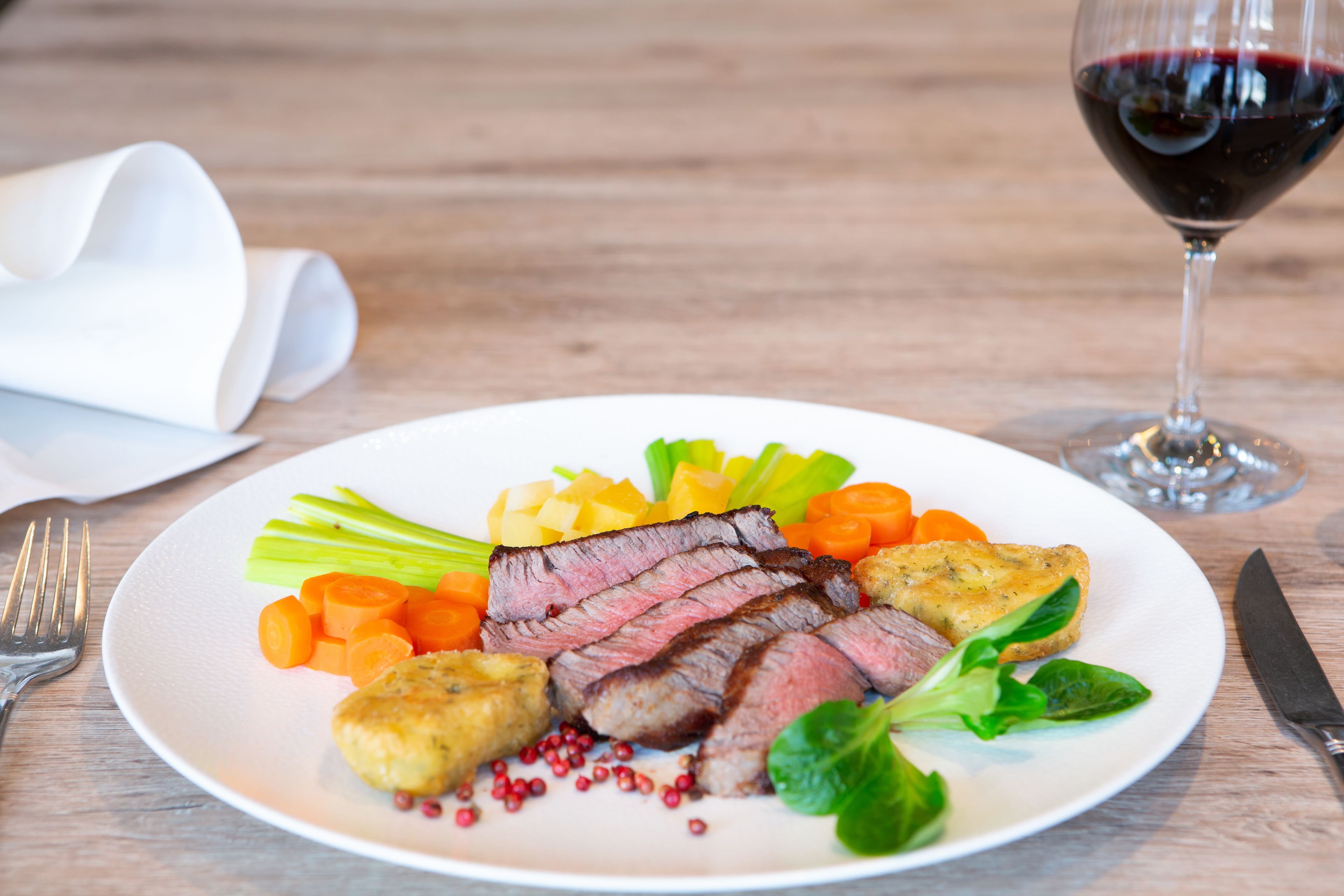 Een bord met plakjes steak, groenten en een glas rode wijn op een houten tafel.