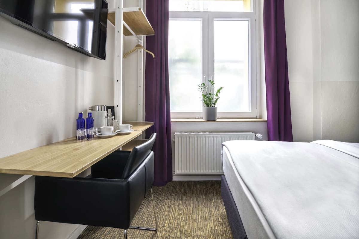 Une chambre d’hôtel avec un lit, un bureau et une chaise, un téléviseur fixé au mur, une fenêtre avec des rideaux violets et une plante en pot sur le rebord de la fenêtre.
