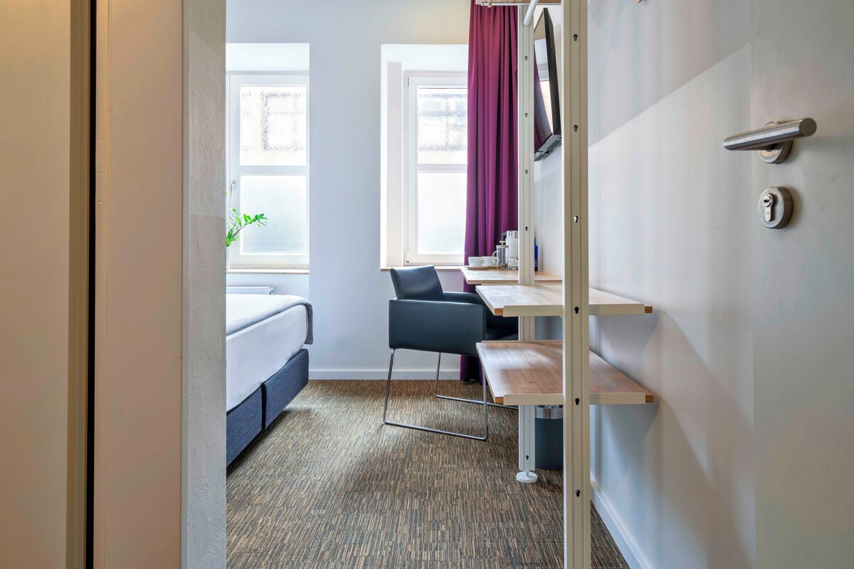 Habitación de hotel pequeña con cama, escritorio con silla, televisor montado en la pared y ventana con cortinas.