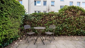 Un pequeño espacio al aire libre con una mesa y dos sillas, rodeado de abundante vegetación y un edificio de fondo.