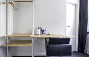 Una habitación pequeña y moderna con un escritorio de madera, una silla negra, una cafetera y una taza.