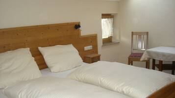 Ein Doppelbett mit Holz-Kopfteil, ein Nachttisch, ein kleines Fenster mit Spitzenvorhängen und ein Stuhl mit Tisch.
