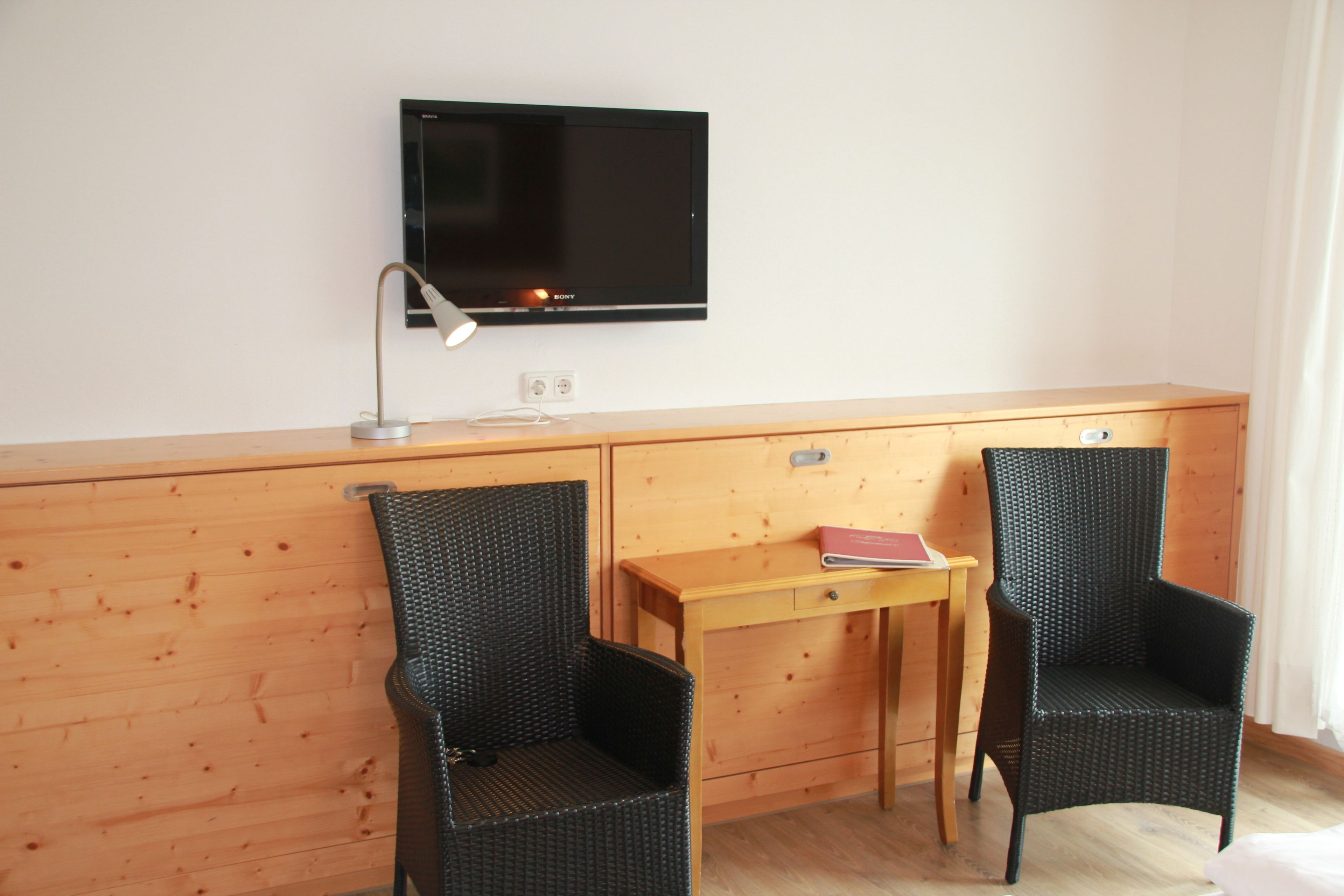 A hotel room with a wooden desk, two wicker chairs, a television, and a small table with a book.
