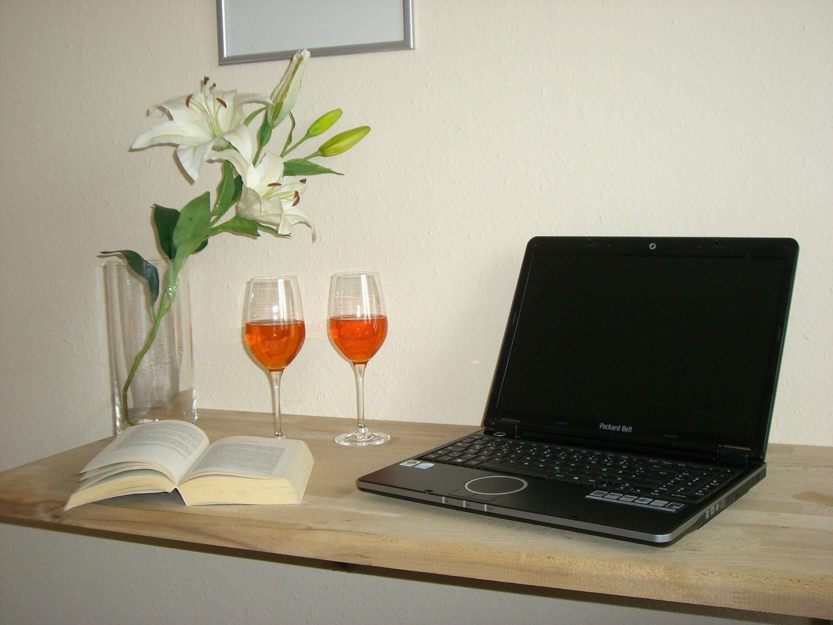 A laptop on a wooden table with two wine glasses and a book beside it.