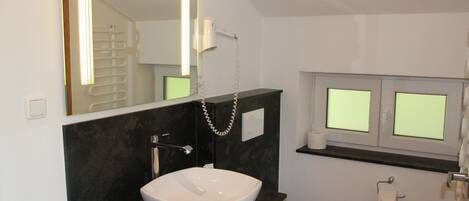 A modern bathroom with a white sink, black countertop, and a large mirror. There is a toilet and a window above it.