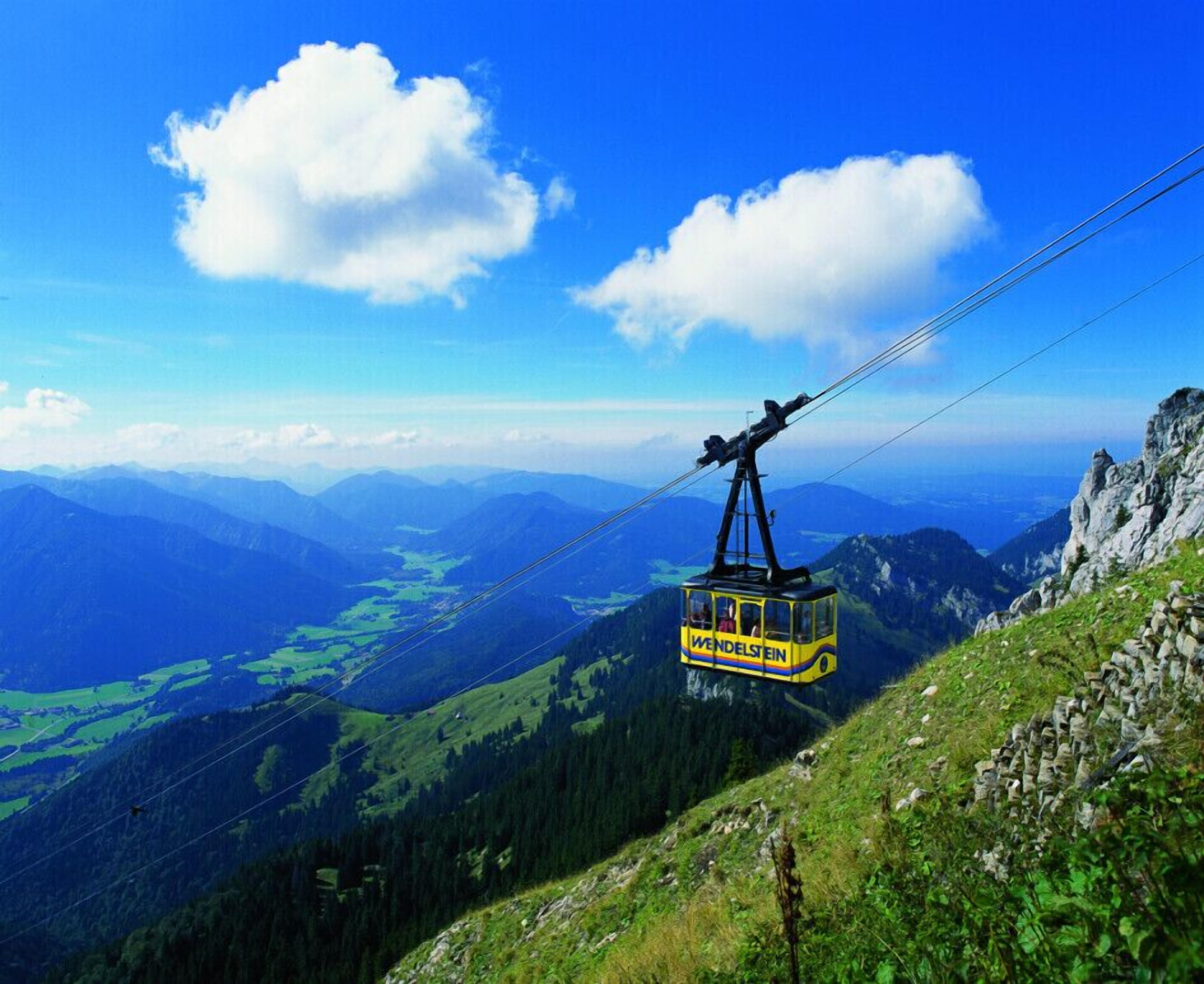 Una funivia con la scritta "Wendelstein" sospesa tramite cavi, che offre una vista su un paesaggio montuoso con valli e boschi.
