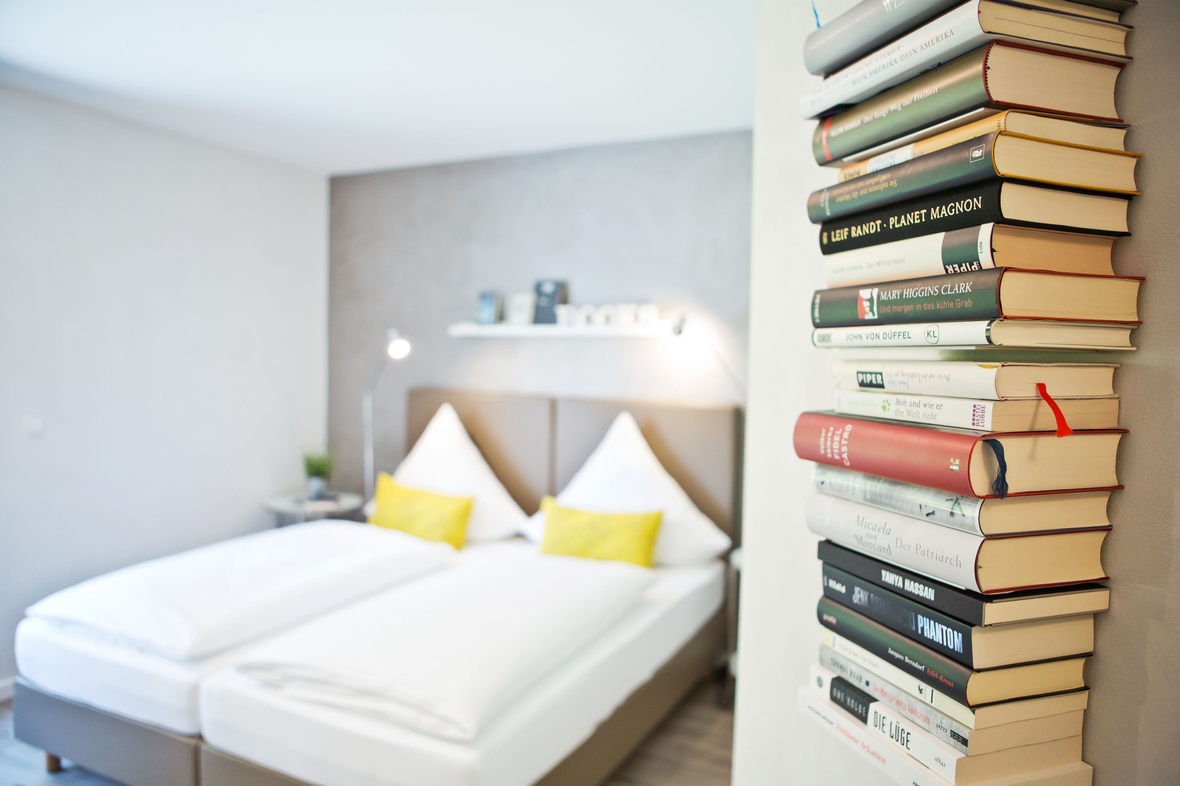 A neatly made bed with white linens and two pillows, a bedside table with a lamp, and a stack of books on a shelf.