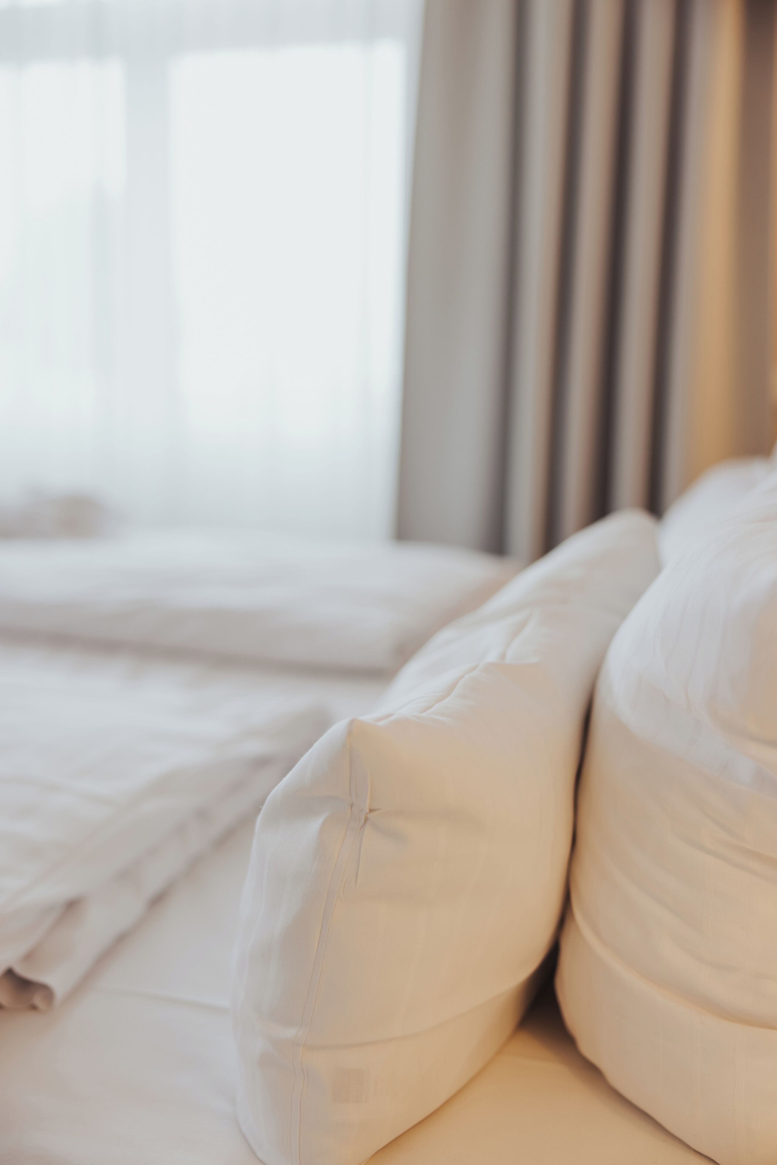 Een netjes opgemaakt bed met witte lakens en kussens, een raam met gordijnen en uitzicht op buiten.